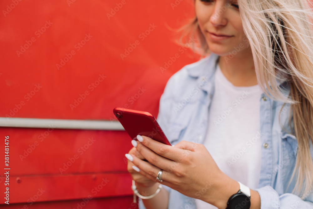 Blond woman texting message on mobile phone against red background outdoors. Copy space. Blond woman texting message on mobile phone against red background outdoors. Copy space.