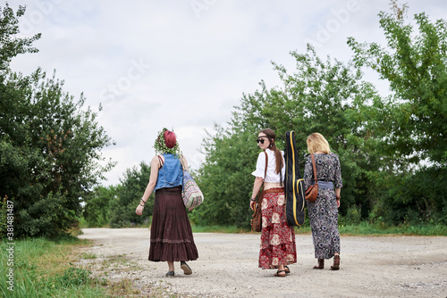 Three hippie women, wearing boho style clothes, holding guitar, walking on dirt road in countryside. Friends, traveling together in summer. Freedom and happiness concept.