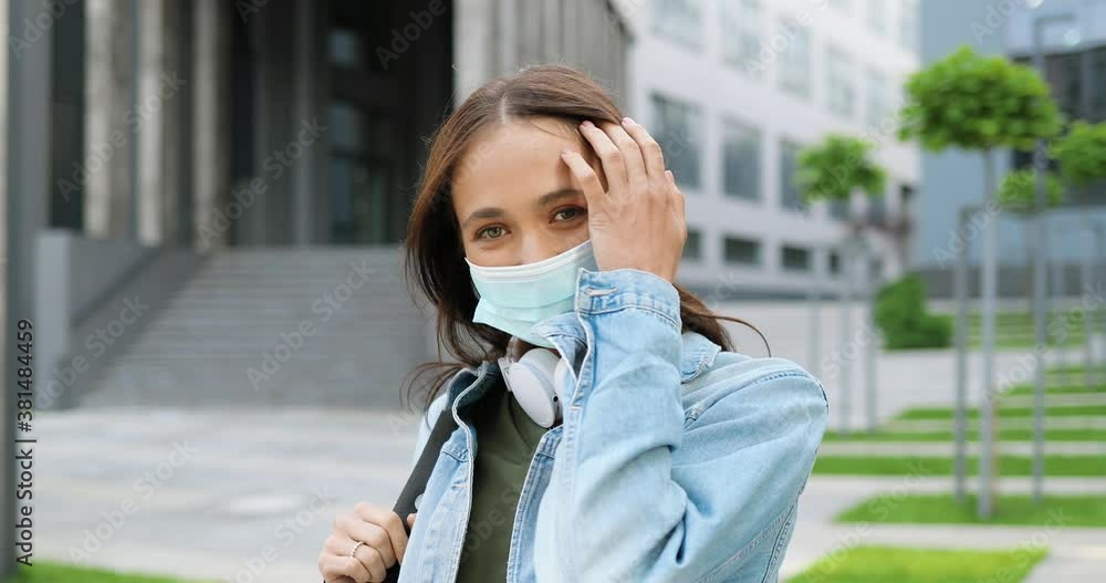 Portrait of young Caucasian beautiful woman in medical mask looking to camera outdoors and laughing. Attractive cheerful female student in headphones and with back at street in quarantine. Pandemic. Portrait of young Caucasian beautiful woman in medical mask looking to camera outdoors and laughing. Attractive cheerful female student in headphones and with back at street in quarantine. Pandemic.