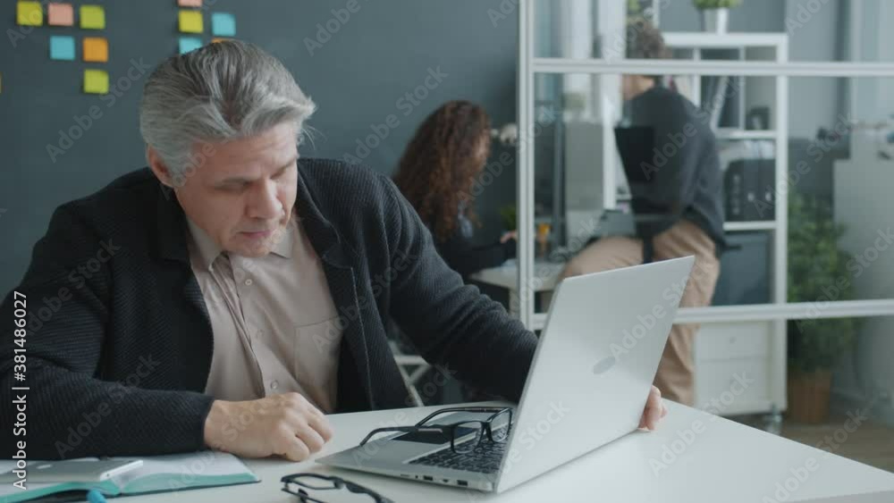 Exhausted office worker mature man is sleeping on desk then waking up and working with laptop typing, female colleagues are working in background. Exhausted office worker mature man is sleeping on desk then waking up and working with laptop typing, female colleagues are working in background.