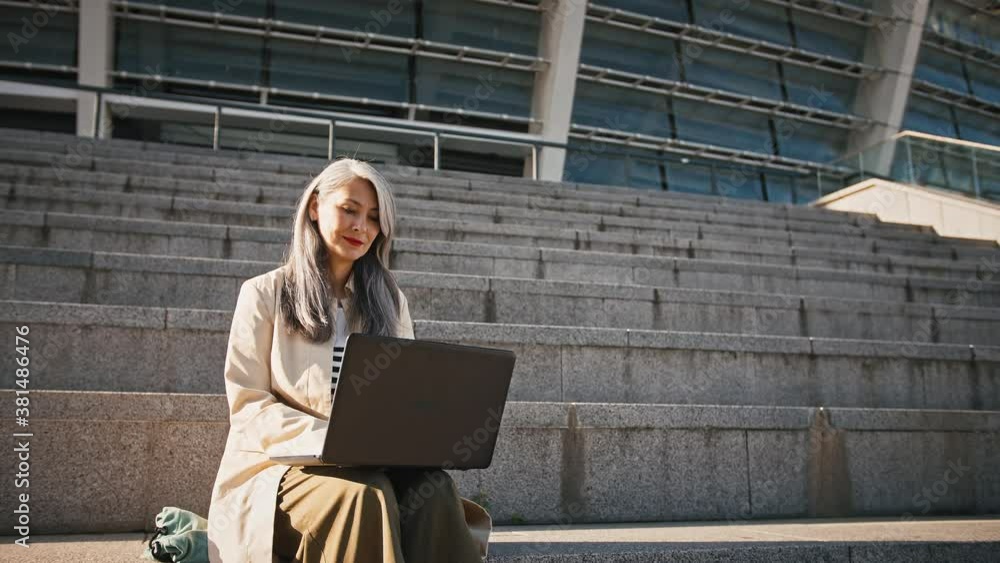 Asian middle-aged woman is working online, typing on laptop computer while sitting on stairs near a stadium Asian middle-aged woman is working online, typing on laptop computer while sitting on stairs near a stadium