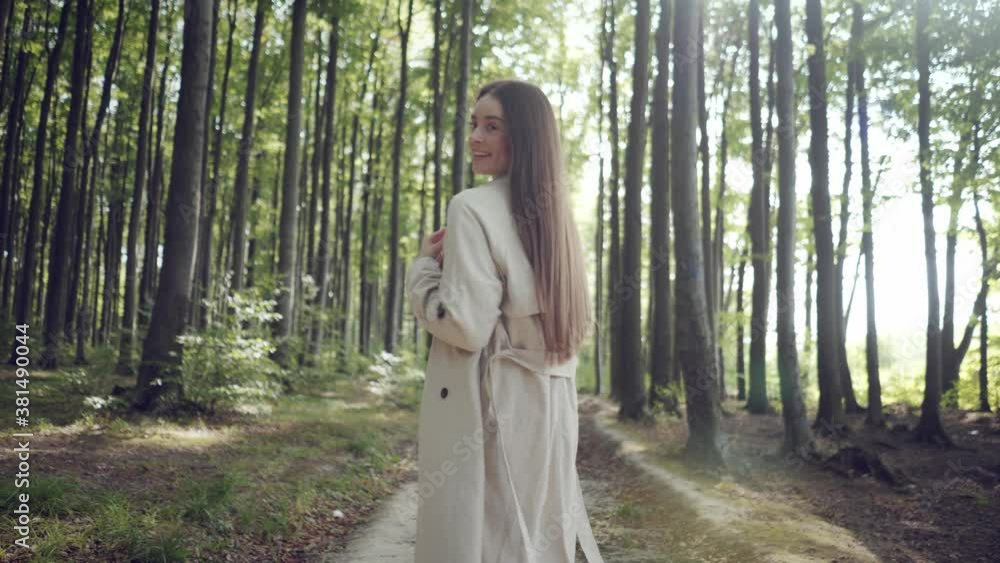 Stylish young brunette woman with red umbrella walking along the path in park or forest area, turning around, smiling, looking at camera. Warm sunny day. Stylish young brunette woman with red umbrella walking along the path in park or forest area, turning around, smiling, looking at camera. Warm sunny day.
