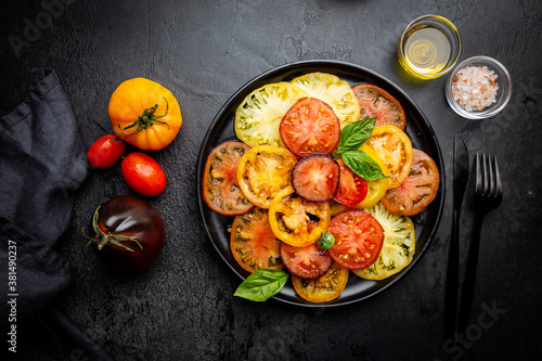 Ripe fresh colorful tomatoes salad with olive oil and basil leaves on black background, top view