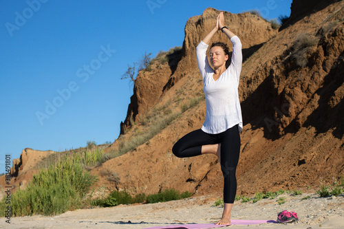 Young woman practicing yoga standing in Vriksha asana with namaste gesture overhead, tree pose on beach, on sport mat and meditating with closed eyes against background of sandy cliffs and blue sky photo