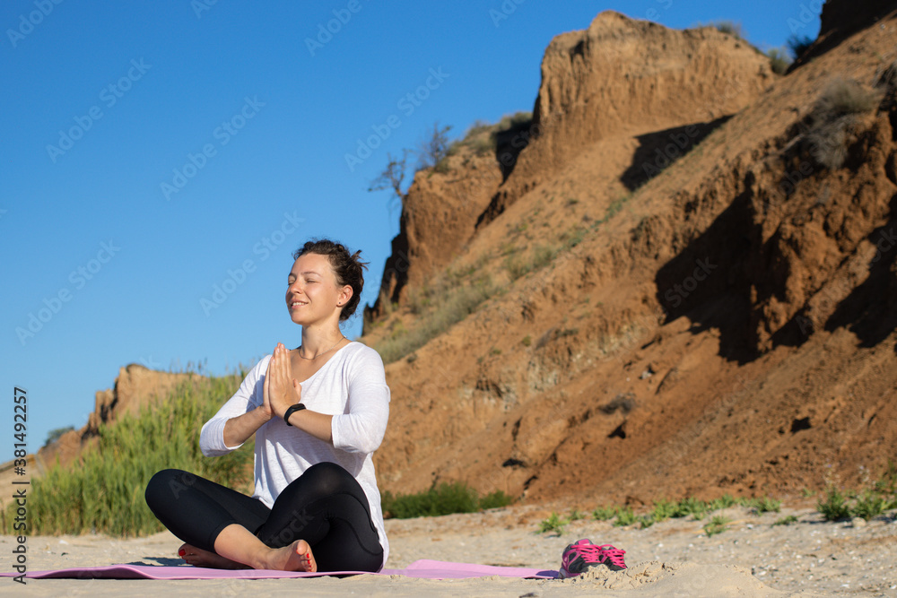 Young beautiful caucasian woman with closed eyes, smiling practicing yoga on the beach, sitting in lotus position asana on mat and meditating against the background of sandy cliffs and blue sky