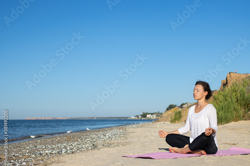 Young beautiful brunette woman practicing yoga, sitting alone in lotus position asana and meditating with closed eyes at seashore in the summer morning. Relax, wellness, healthy lifestyle. Copy space