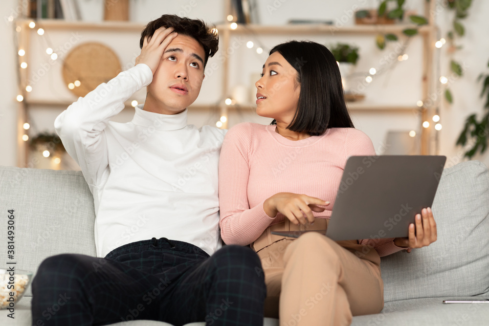 Asian Wife Shopping Online With Shocked Husband Sitting At Home Asian Wife Shopping Online With Shocked Husband Sitting At Home