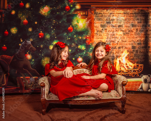 Christmas portrait of two little smiling girls wearing red costumes on Christmas tree background sitting on a vintage bench © iryf