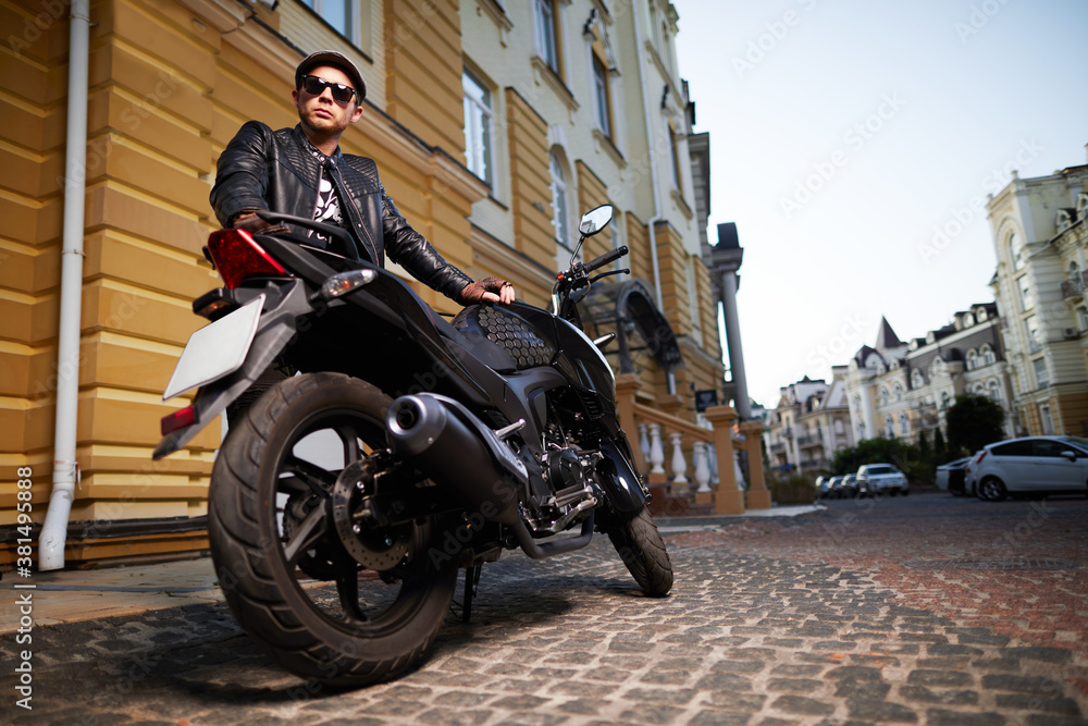 A handsome man in sunglasses and leather jacket standing near motorbike on the street.
