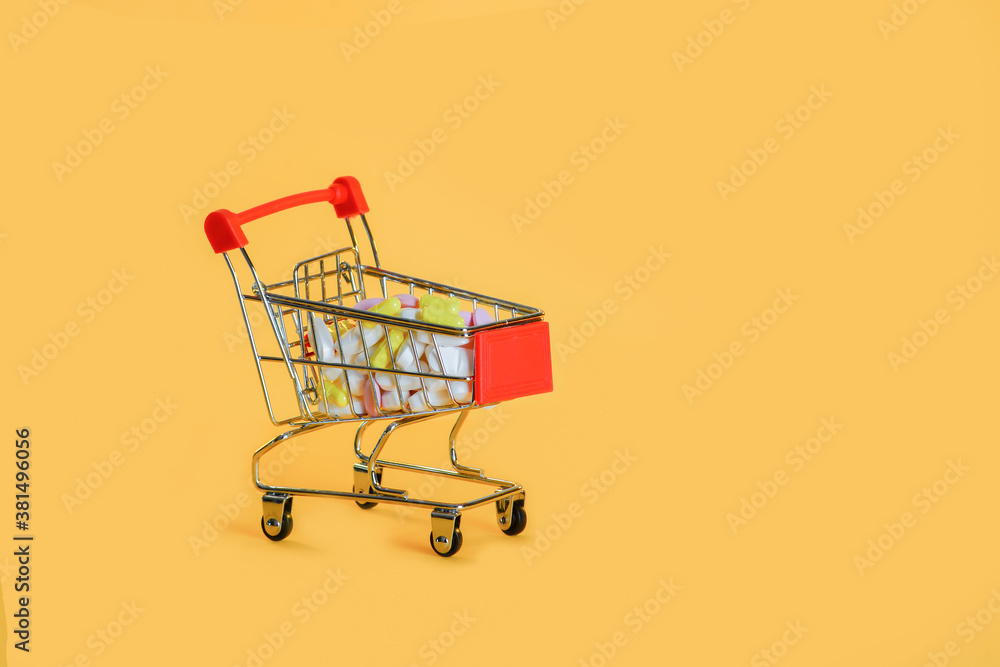 Small grocery cart with a bunch of white, pink and yellow pills,isolated on yellow background.Purchase of medicine Small grocery cart with a bunch of white, pink and yellow pills,isolated on yellow background.Purchase of medicine