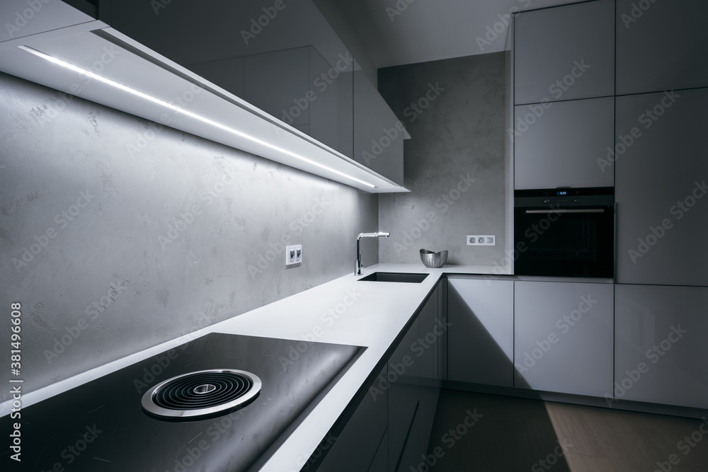 Modern minimal kitchen with white worktop,bright light strip turned on and modern appliances.Focused on hob with integrated extractor hood, then gradually blurs in background(out of focus) direction. - obrazy, fototapety, plakaty 