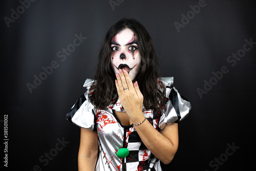woman in a halloween clown costume over isolated black background with her hands over her mouth and surprised, looking side