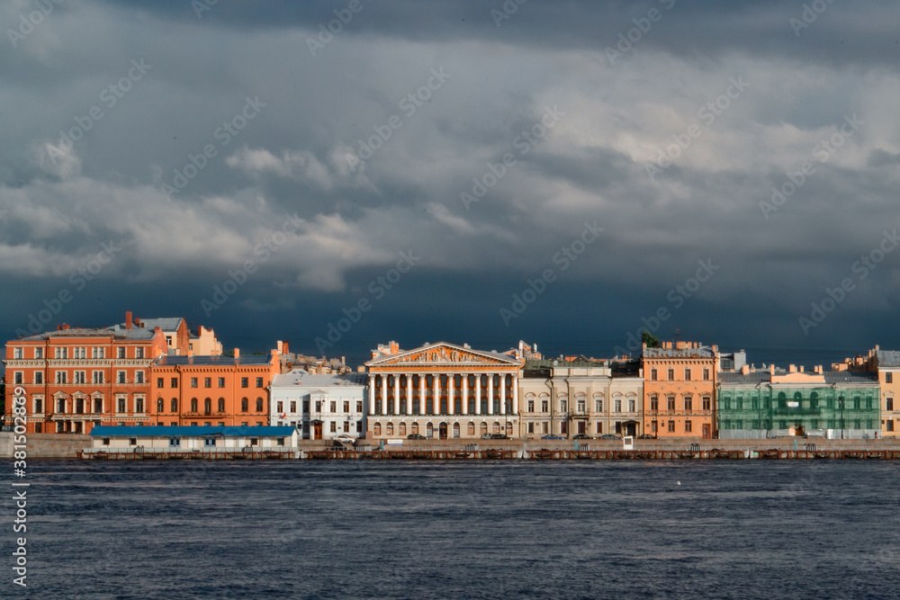 Embankment with colorful bright buildings on banks of blue Neva River in the light of setting sun. Saint Petersburg, Russia. Dark clouds.