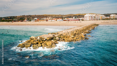 Rocky pier or breakwater on the sandy beach. Beautiful green blue ocean with surfers. Aerial view or drone photo. Costa Da Caparica, Setubal, Almada, Portugal.  photo