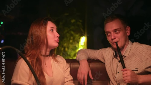 Cute couple in love relaxing on a date. Media. Caucasian young man and woman in black smoking a hookah while sitting at the table in cafe indoors together. photo