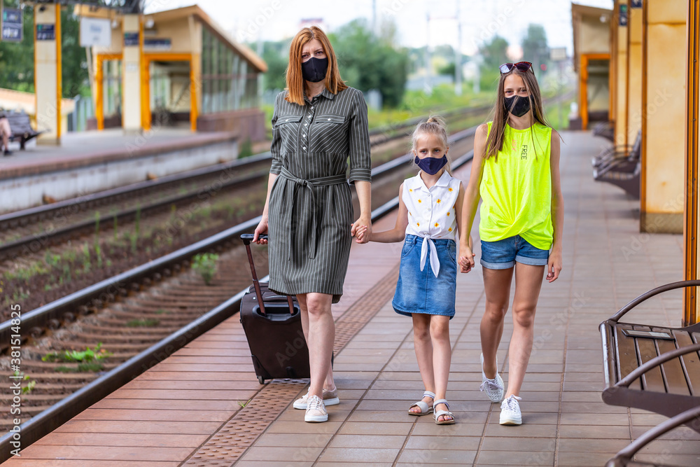 Fototapeta premium Safe family travel. Mother and two daughters in face masks are waiting for take off by train at the railway station. City view.