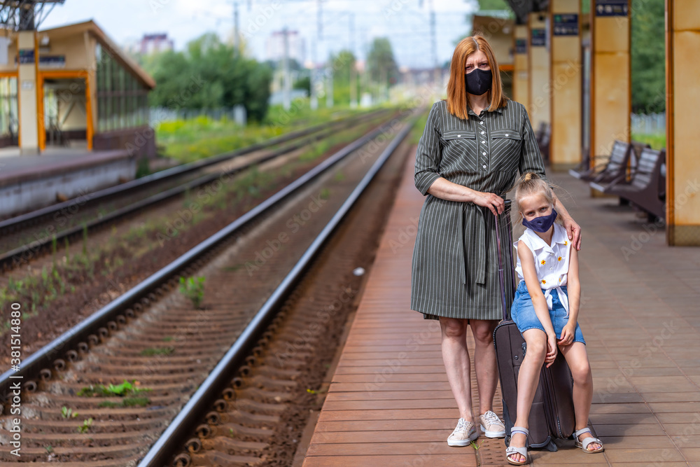 Fototapeta premium Caucasian female with daughter in a black face masks with luggage are waiting for take off at the railway station. Safe travel.