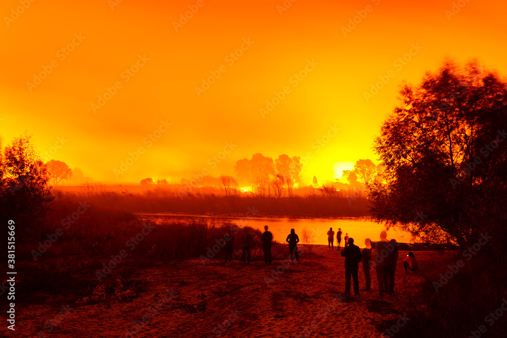 People near the river look at burning fields and forest