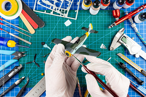 desktop view from above of assembly and painting of retro scale model fighter plane concept background. modeling tools airbrush gun paint kit parts blue green cutting mat knife and brush work desk