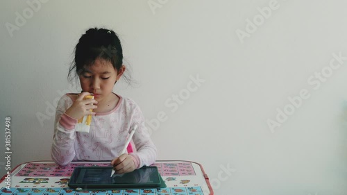 Portrait of little Asian girl drinking milk while using tablet, sitting at desk and doing homework isolated on white background. E-learning and education concept.