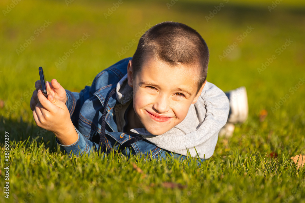 A cheerful boy in a denim jacket lies on a green lawn and plays with a smartphone in a city park. Concept of the problem of children's passion for mobile games and cartoons A cheerful boy in a denim jacket lies on a green lawn and plays with a smartphone in a city park. Concept of the problem of children's passion for mobile games and cartoons