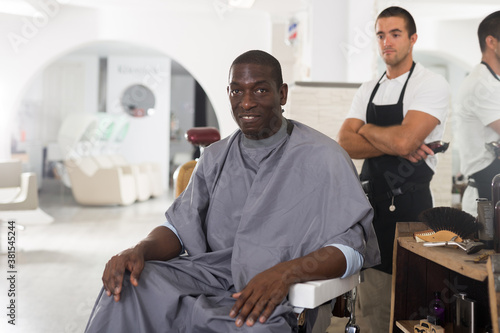 Portrait of happy African-American male client of barbershop after haircut performed by skilled hairstylist