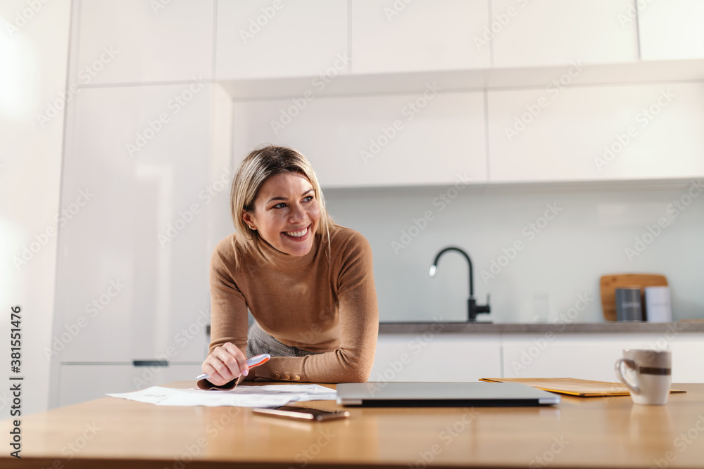 Young gorgeous blond businesswoman leaning on kitchen table and filling up bills while standing in kitchen at home. Young gorgeous blond businesswoman leaning on kitchen table and filling up bills while standing in kitchen at home.