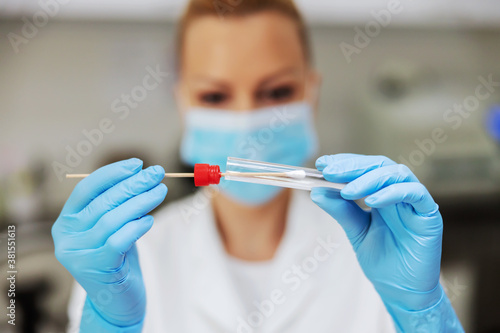 Blond female lab assistant in sterile uniform with rubber gloves and surgical mask on holding cotton swab. Laboratory interior.