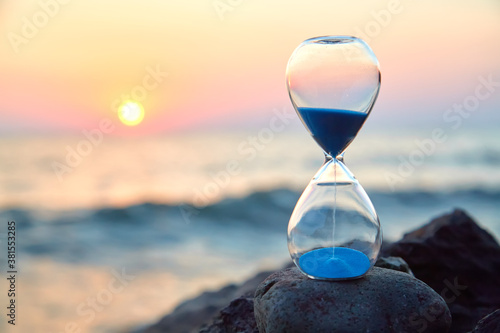 Hourglass with sand standing on rock. Sunset over sea and nature landscape. Running of time and relax idea, copy space