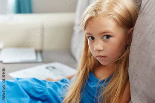 portrait of caucasian child girl in dress sitting on sofa in living , beautiful kid girl is sad. mom and dad drawing in the background as a symbol of divorce close up sad portrait photo