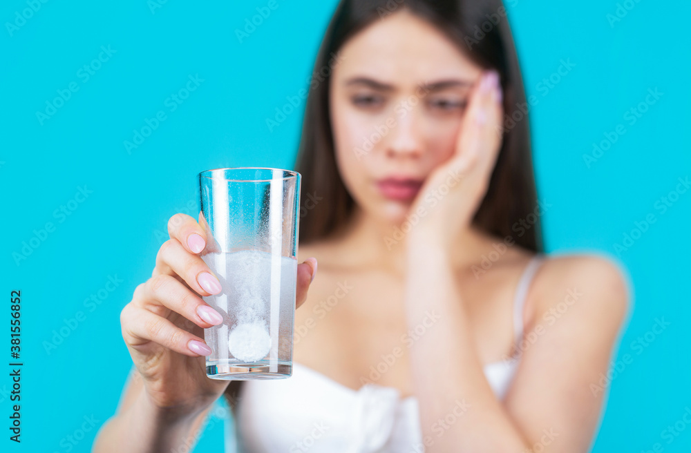 Woman taking drugs to releave headache. Brunette take some pills, holds glass of water, isolated on blue. Young woman taking pill against headache. Brunette taking a pill with a glass of water Woman taking drugs to releave headache. Brunette take some pills, holds glass of water, isolated on blue. Young woman taking pill against headache. Brunette taking a pill with a glass of water