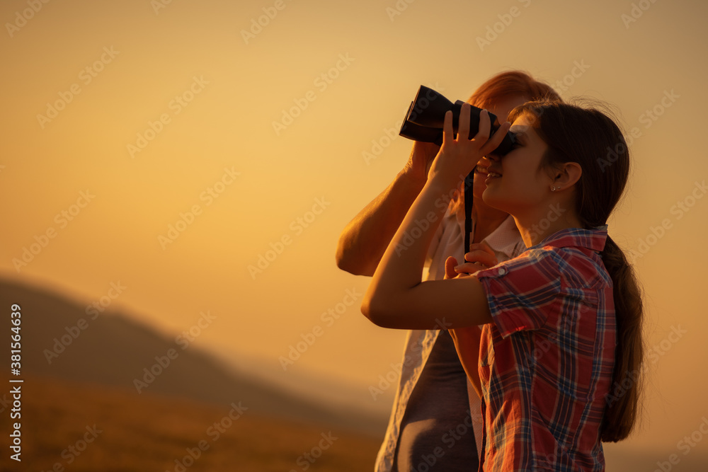Happy grandmother and granddaughter are using binoculars in nature in sunset.
