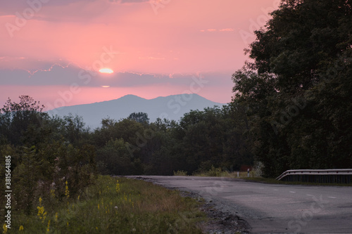 Country road at sunrise. The sun rises against the background of the mountains. Green grass and a road stretching into the distance.