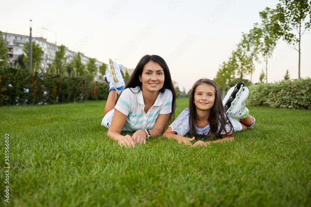mother and daughter in roller skates are sitting on grass and looking at the camera in summer park. mother and daughter in roller skates are sitting on grass and looking at the camera in summer park.