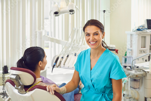 Smiling woman dentist at her office near the patient in the dental chair looking at the camera.