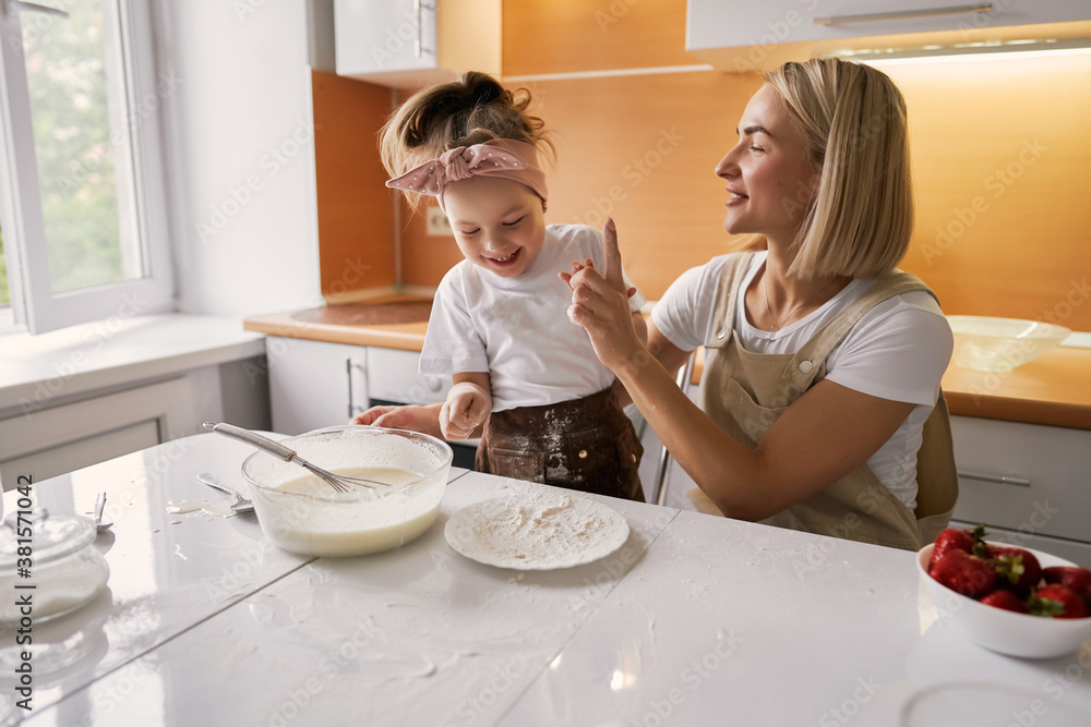 happy mother baking with little daughter in apron and cook hat working with flour , bowl and spoon preparing dough teaching the kid baking and having fun together. happy mother baking with little daughter in apron and cook hat working with flour , bowl and spoon preparing dough teaching the kid baking and having fun together.