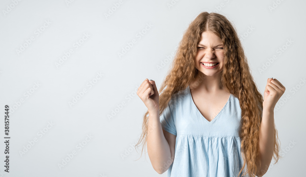 Joyful girl keeps fists clenched and exclaims in triumph, celebrates success. Studio shot white background. Copy space