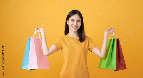 Young smiling asian woman holding multi coloured shopping bags on light yellow background.