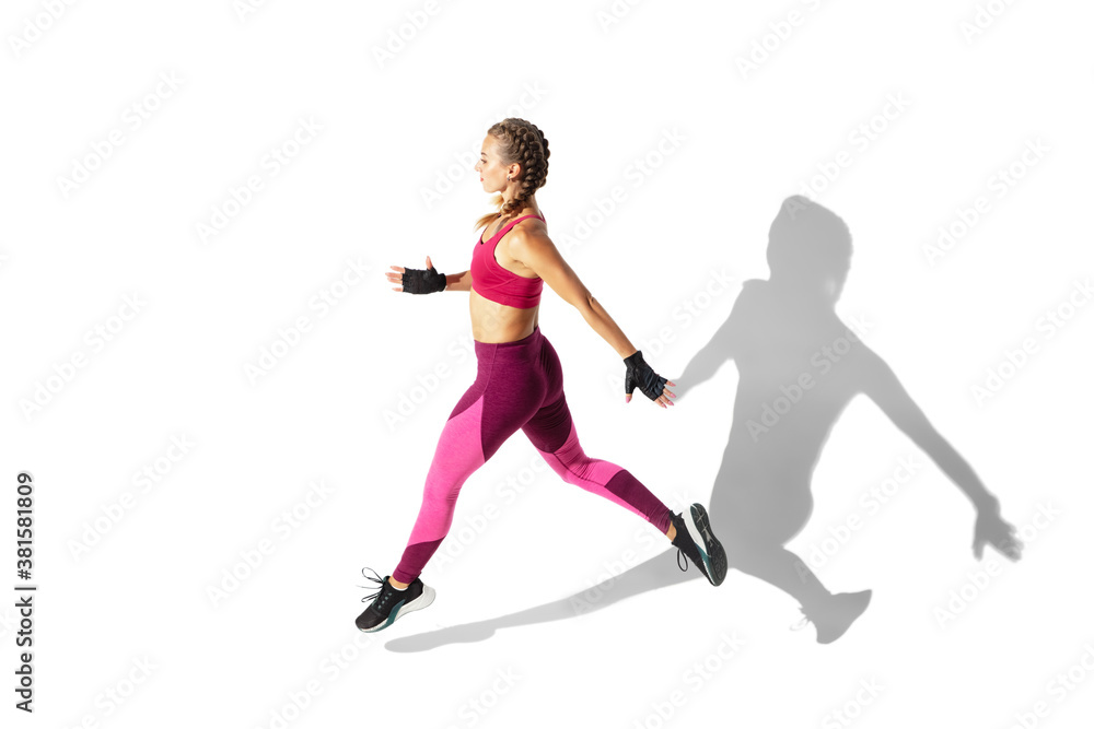Running. Beautiful young female athlete practicing on white studio background, portrait with shadows. Sportive fit model in motion and action. Body building, healthy lifestyle, style concept.