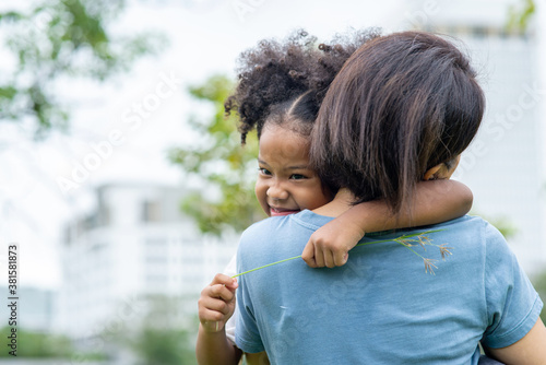 Happy affectionate mixed race family. Cute little child girl embrace her mother with toothy smile. Mom carrying daughter and spending time together in summer outdoor weekend holiday vacation.