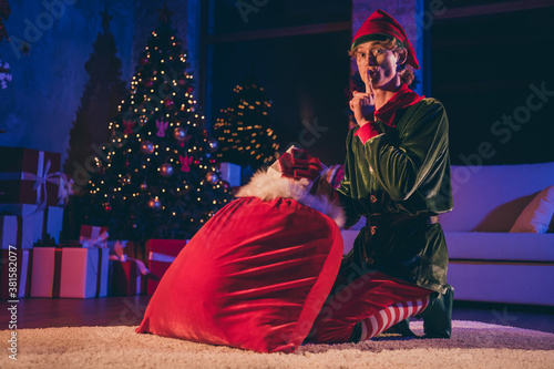 Full body photo of elf sit floor with big sack x-mas christmas delivery put finger mouth wear green cap in house indoors evening lights photo