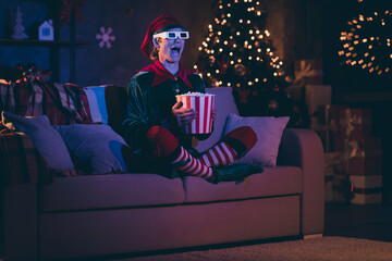 Full size photo of amazed elf sit couch watch three dimensional film with pop corn box in evening christmas x-mas decor house indoors