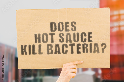 The question " Does hot sauce kill bacteria? " on a banner in men's hand with blurred background. Dangerous. Hazardous. Germs. Cooking. Spicy