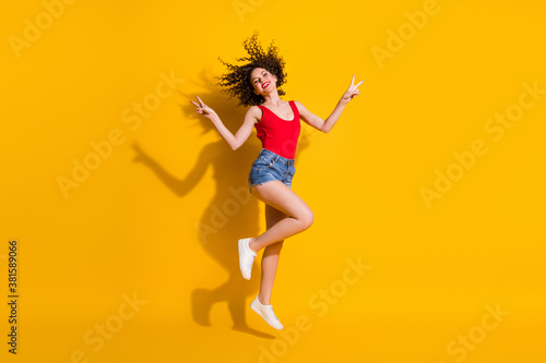 Full length body size view of her she nice attractive slim fit cheerful cheery optimistic wavy-haired girl jumping having fun showing v-sign isolated bright vivid shine vibrant yellow color background