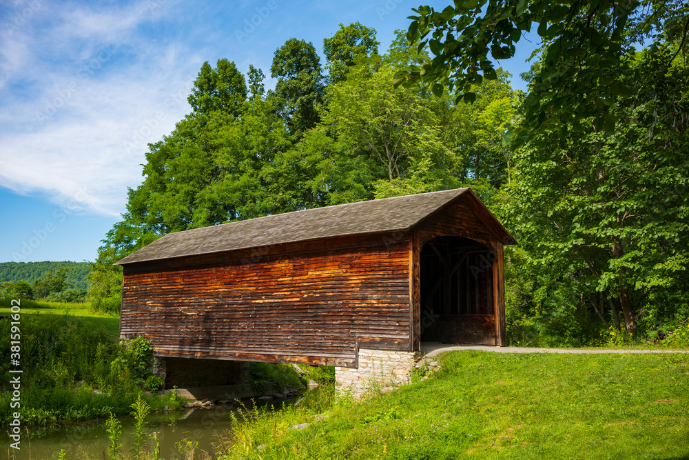 The Hyde Hall Covered Bridge, built in. 1825 and is the oldest existing covered bridge in the U.S., rests at the end of a dirt road during a summer day. 