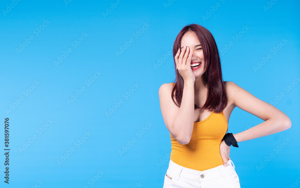 Fototapeta premium Cheerful young woman brown long hair wear short jeans holding hand on her face while standing over isolated blue background. Positive girl smiling posture while looking at camera with copy space.