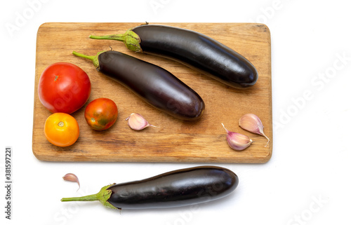 Three eggplants, tomatoes and cloves of garlic on a wooden kitchen board on a white background. Ingredient for fitting a dietary vegetable dish.
