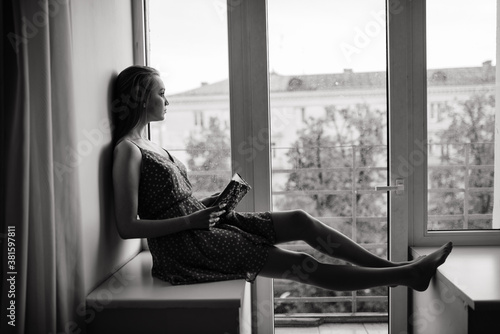 Pensive dreamy female holding book at home looking at big window dreaming, thoughtful young woman