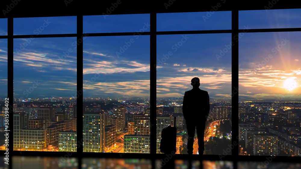 The male with suitcase standing near the panoramic window with a sunset