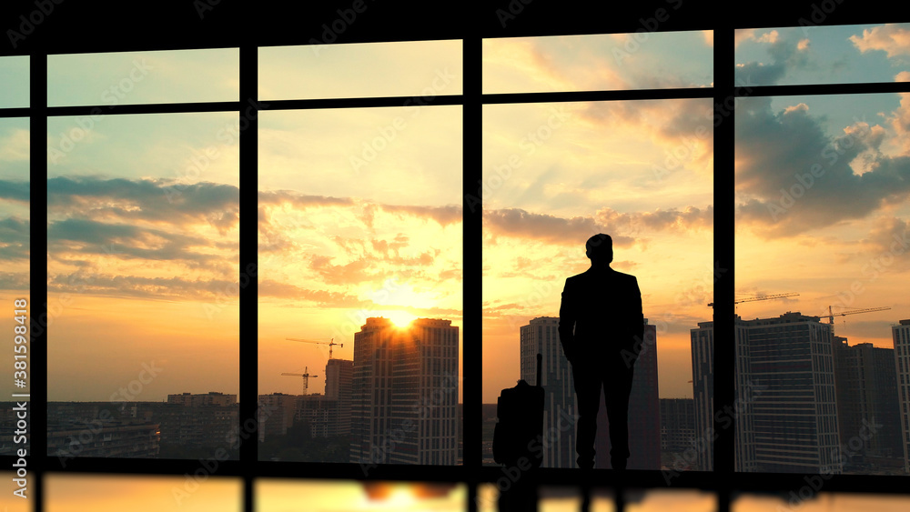 The man with a suitcase standing near a panoramic window against a sunset The man with a suitcase standing near a panoramic window against a sunset